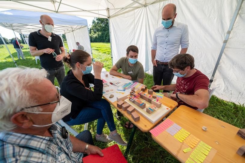 Menschen mit Maske gruppiert um einen Tisch mit einem Holz-Baublock-Modell