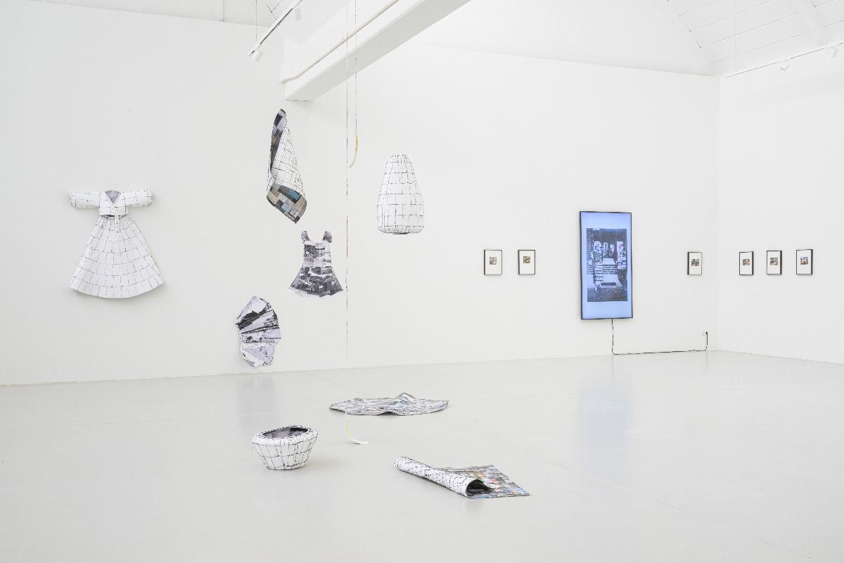 Ausstellungsansicht der Installtion "Connected Spaces" von Gisoo Kim. Mehrere Objekte, die aus zuammengenähten Fotografien bestehen und Gestalt annehmen, etwa ein Kleid, Vasen, Kokons. Im Hintergrund die Virdeoinstallation von Lily Lulay