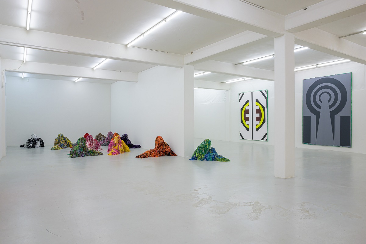 Ausstellungsansicht mit großen Malereien die an Wappen oder Symbole erinnern von Pia Rosa Dobrowitz und kauernde Figuren in bunten Tarnanzügen von Yongkuk Ko