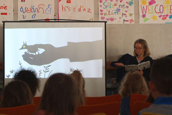 Eine Frau liest aus einem Buch vor, daneben eine Leinwand mit Schattenspiel