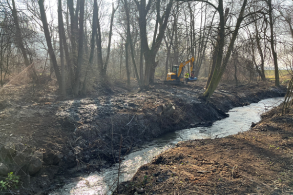 Wassergraben in Erde, kahle Bäume und ein Schaufelbagger im Hintergrund
