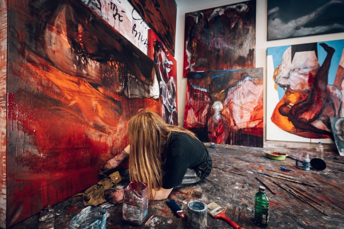 Person sitz in einem Atelier mit vielen Bildern und malt gerade an einem.