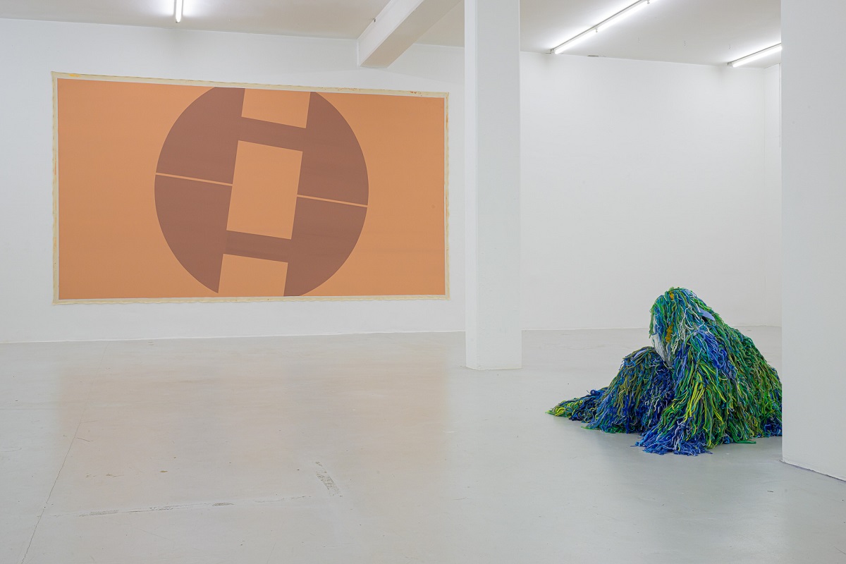 Ausstellungsansicht mit einer großen Malerei die an ein Smbol erinnert von Pia Rosa Dobrowitz und eine kauernde Figur in grün-blauen Tarnanzug von Yongkuk Ko