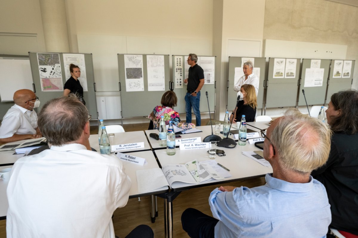 Der Gestaltungsbeirat sitzt in einem großen Raum verteilt, an einer Wand hängen mehrere Baupläne.