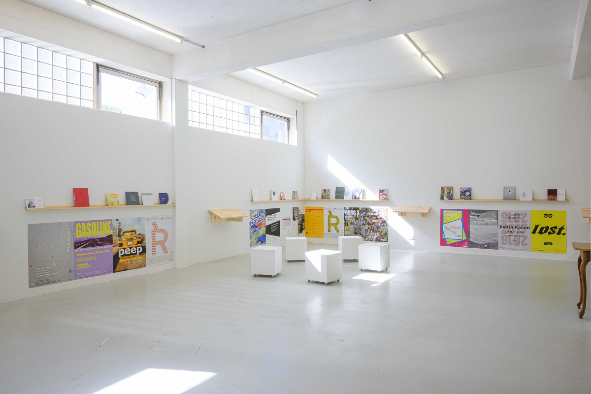 Ausstellungsansicht von "Abschied vom L6". Über Plakaten von vergangenen Ausstellungen befinden sich schamle Regale auf denen von Künstler_innen mitgebrachte Kataloge präsentiert werden