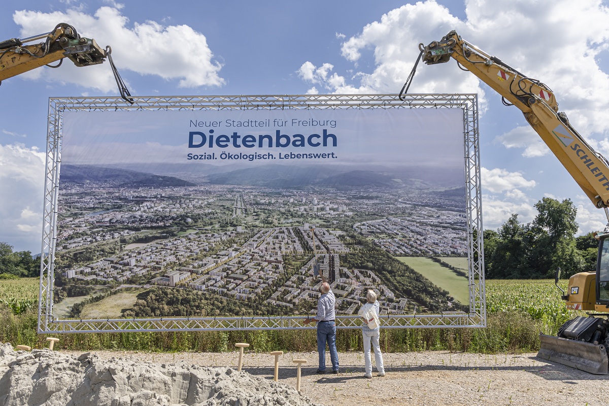 Zwei Bagger halten eine Leinwand hoch, auf der das neue Stadtgebiet Dietenbach abgebildet ist.