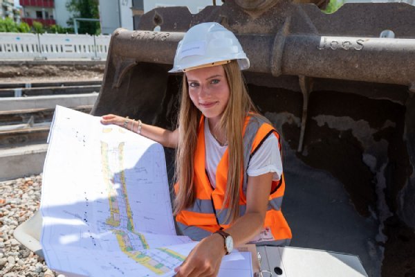 junge Frau mit Bauhelm sitz auf einer Baggerschaufel und liest einen Bauplan