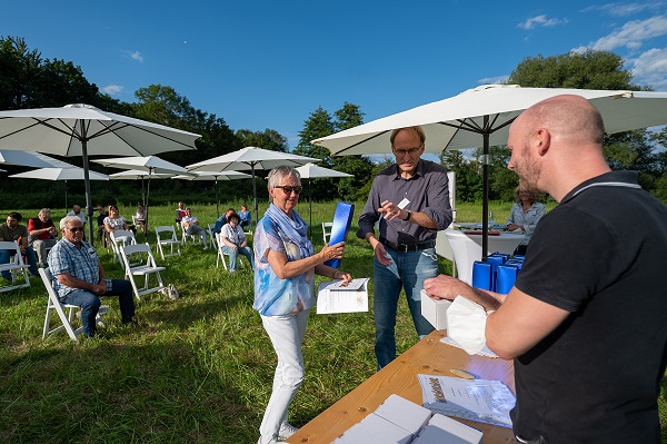 Eine Dame mit Sonnenbrille und zwei Männer übergeben sich Geschenke auf einer Wiese unter blauem Himmel