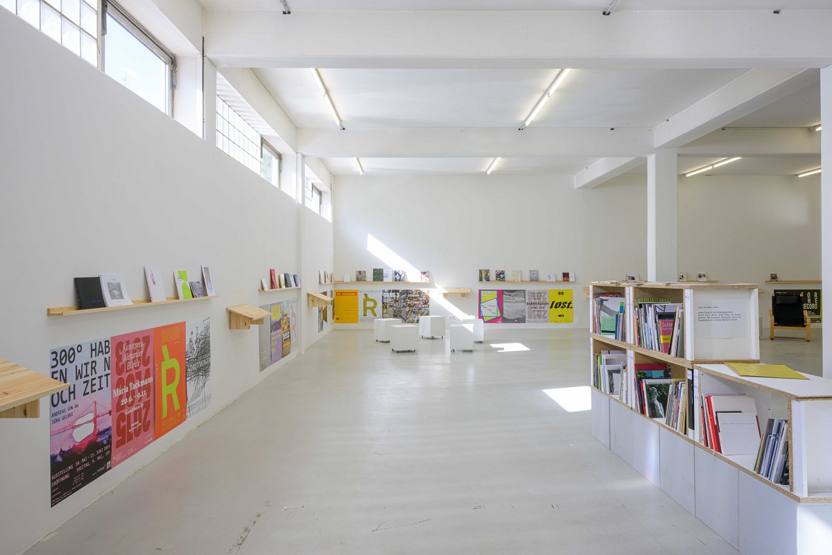 Ausstellungsansicht von "Abschied vom L6". Über Plakaten von vergangenen Ausstellungen befinden sich schamle Regale auf denen von Künstler_innen mitgebrachte Kataloge präsentiert werden. 