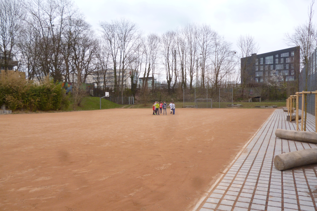 Großer Hartplatz, von Bäumen und Gebäuden umringt. In der Mitte haben Kinder in Fußballklamotten einen Kreis gebildet.