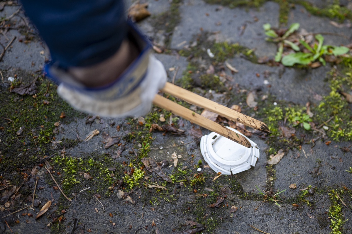 Eine Hand mit einem Handschuh und einer Holzzange hebt gerade einen Kaffeebecherdeckel aus Plastik auf.