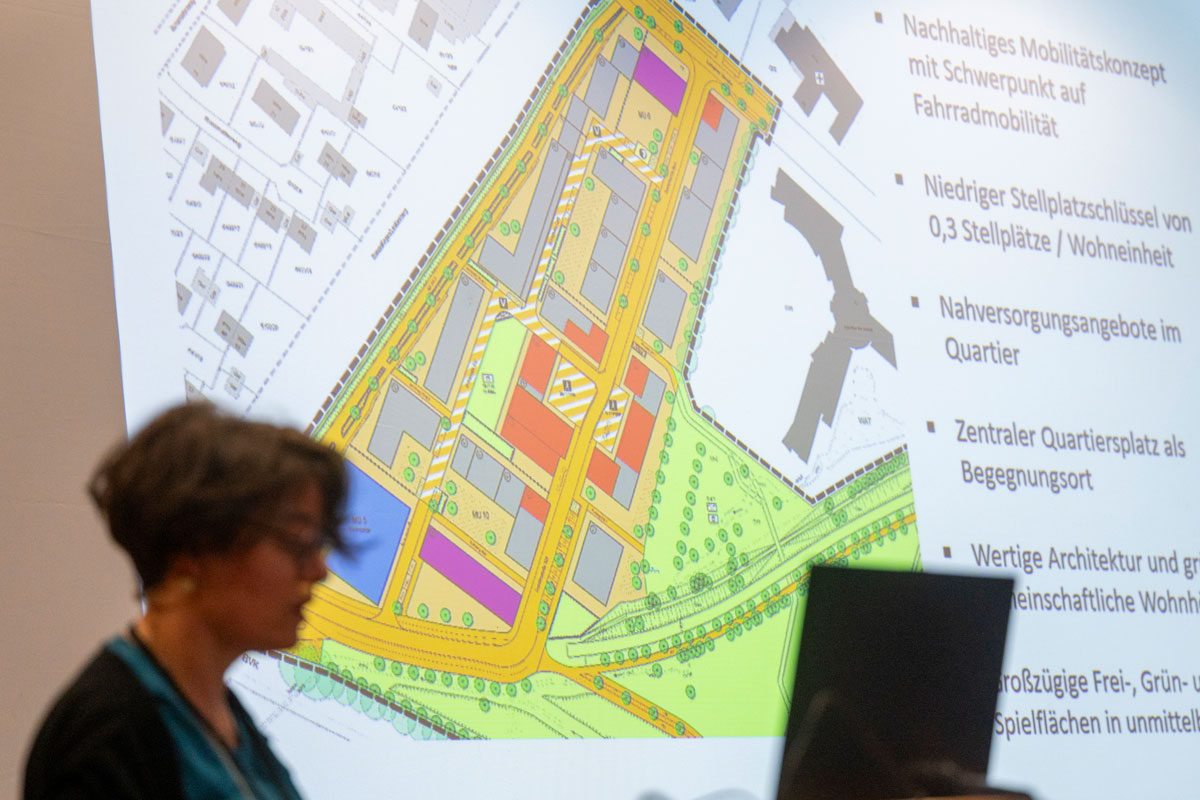 Person erklärt vor Leinwand etwas, im Hintergrund ein Bauplan