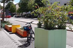 Kastenmöbel aus Holz und Kunststoff zum Sitzen, stehen auf einem Parkplatz, im Vordergrund ein Pflanzenkübel
