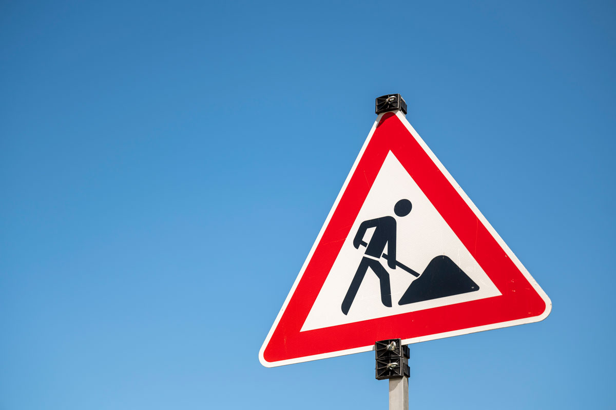 Baustellenschild vor blauem Himmel zeigt Bauarbeiter*in mit Schaufel